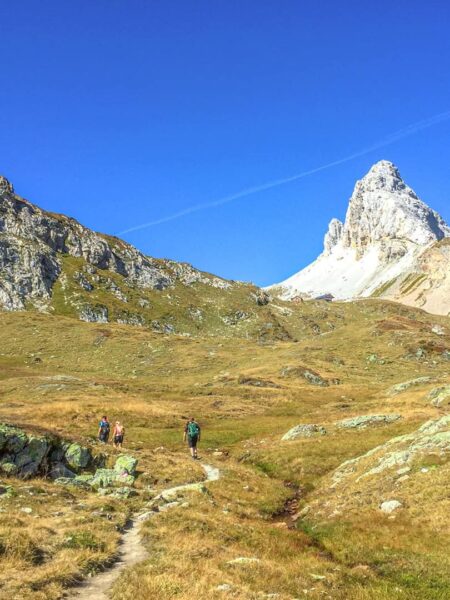 Drei Personen in Wanderkluft marschieren über einen schmalen Weg einen Hang hinauf durch das Lesachtal. Sie verlassen das Bild nach links, im Hintergrund sieht man den spitzen Gipfel eines Berges.