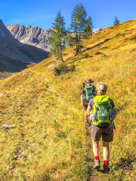 Zwei Personen in kurzen Hosen Wandern mit ihren Rucksäcken einen Hang in Kärnten hinauf. Die Sonne scheint hell an dem Herbsttag, an dem sie gerade unterwegs sind, im Hintergrund sieht man karge Berge und vereinzelte Bäume.