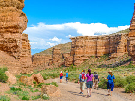 Eine kleine Gruppe auf Wanderreise geht durch einen Canyon in Kasachstan. Sie tragen Rucksäcke, rund um sie herum erheben sich große Formationen aus rotem Fels in den Himmel.