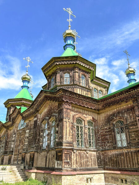Das Bild zeigt eine russisch-orthodoxe Kirche in Karakol in Kirgistan. Sie ist gebaut aus stark verziertem Holz und wiest grünliche Spitze Türmchen auf, die sich vom blauen Himmel abheben.