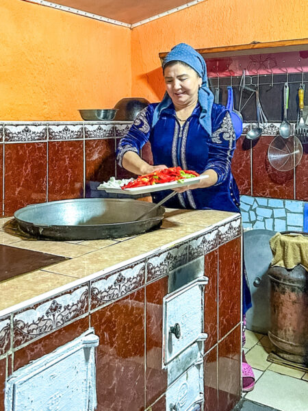 Eine Frau einer uigurischen Familie steht in einer Küche und schüttet gerade Gemüse in einer große Pfanne oder einen großen Topf. Sie lacht in die Kamera.