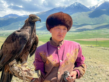 Ein Adler sitzt auf der Hand eines zentralasiatischen Adlerjägers. Der Vogel wirkt ruhig, während der Mann der Gruppe hinter der Kamera etwas erklärt. Hinter ihm kann man die weitläufigen Steppen, sowie hohen Berge Kirgistans erkennen.