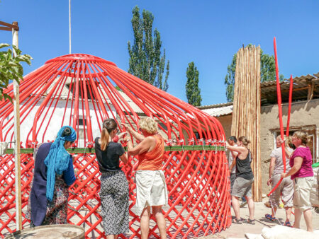 Mehrere Personen auf Wanderreise arbeiten zusammen daran, eine traditionelle Jurte aus roten Holzstäben zu errichten.