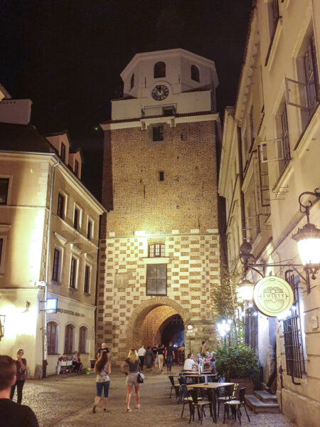 Das Foto zeigt den trinitarischen Turm in Lublin bei Nacht. Im Vordergrund kann man das Schild einer Bar erkennen, sowie mehrere Personen auf Wanderreise, die auf den Turm zugehen.