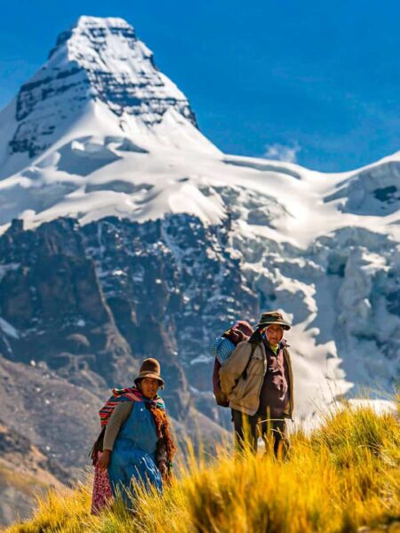 Zwei Menschen aus Bolivien gehen einen Hang im Cordillera Real hinauf. Im Hintergrund ragt ein Berggipfel in den Himmel hinauf und die Sonne strahlt auf die beiden Personen herab.