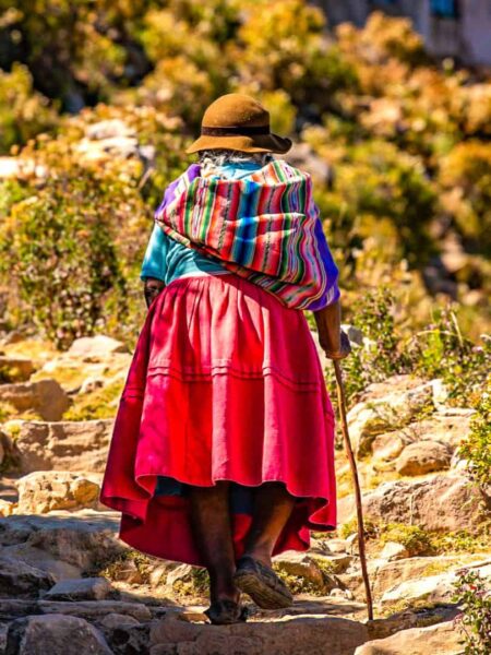 Eine ältere bolivianische Frau geht die Stufen eines Weges auf der Sonneninsel hinauf. Sie trägt einen Hand und traditionell farbenfrohe Kleidung.