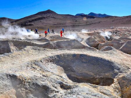 Mehrere Personen in den roten Jacken von Weltweitwandern gehen durch die Salar de Uyuni in Bolivien. Rund um sie befinden sich Geysire, die Dampf in die Luft der großen bolivianischen Salzebene blasen.