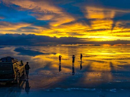 Mehrere Reisende einer kleinen Reisegruppe von Weltweitwandern schauen zusammen mit ihren Guide von einem geparkten Auto aus in den Sonnenuntergrag in der Salar de Uyuni. Das Licht der untergehenden Sonne reflektiert sich auf der leicht feuchten Salzebene.