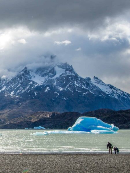 Auf dem Foto kann man den Grey Lake im Torres del Paine Nationalpark in Patagonien sehen. Ein Eisberg treibt auf dem Wasser und zwei Personen auf einer Wanderreise schauen über den See hinweg bis zu einem Berg jenseits des Gewässers.