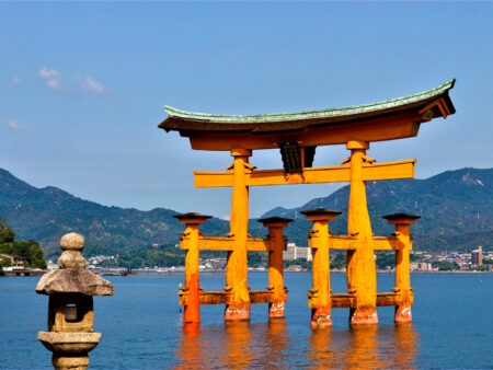 Der rote Torbogen, oder Torii, des UNESCO-Welterbeschreins Itsukushima steht im Fluss auf der Insel Miyajima. Man sieht ihn vom Land aus, wodurch er wie am Wasser zu schwimmen scheint. Im Hintergrund kann man das Bergland Japans erkennen.