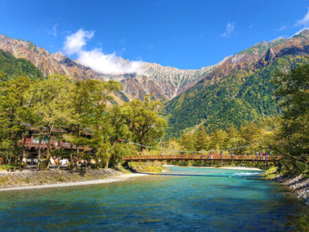 Man sieht eine Brücke über einen Fluss im Tal des Kamikochi. Im Hintergrund erheben sich kahle Berge, während der kristallklare Fluss zu beiden Seiten von dichter Vegetation begrenzt ist.