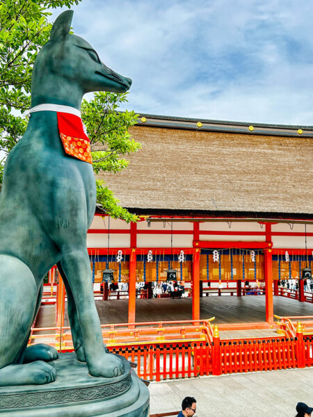 Eine stilisierte Statue eines Fuchses oder Kitsune steht im Schrein Fushimi Inari-Taisha in Japan. Im Hitnergrund sieht man die klassisch roten Gebäude des Schreins.