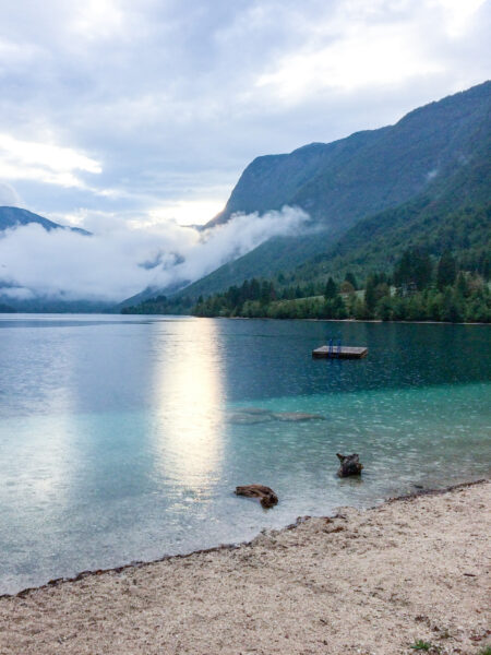 Der Bohinj See in Slowenien unter einem wolkenverhangenden Himmel lädt mit seinem klaren Wasser zum Baden ein. Das Foto wurde von einem Sandstrand aus im Zug einer Wanderreise mit Weltweitwandern aufgenommen.