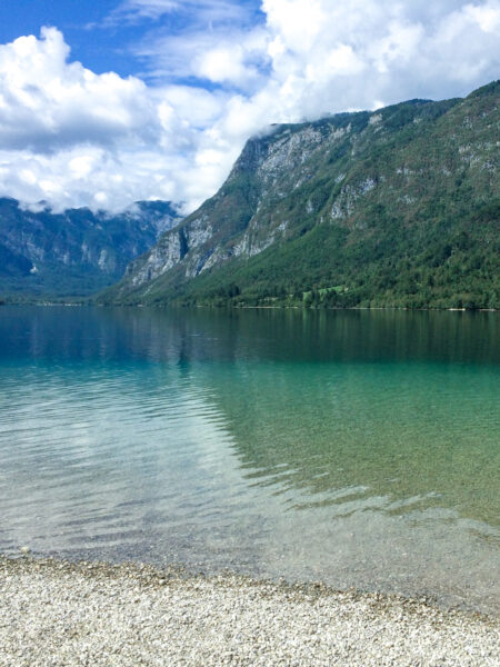 Ein Bild eines Kiestrandes am Bohinj-See in Slowenien. Im Hintergrund sieht man die hohen Berge, die den See umrahmen und zum Wandern einladen.