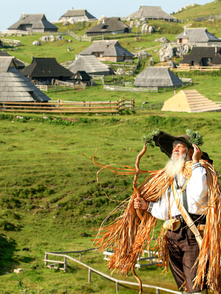 Man sieht einen Einheimischen auf der Velika Planina, einer Hochalm in Slowenien. Hinter ihm kann man mehrere traditionelle Gebäude der Region erkennen, er selbst hält einen langen Wander- oder Hirtenstock und trägt einen breitkrempigen Hut.