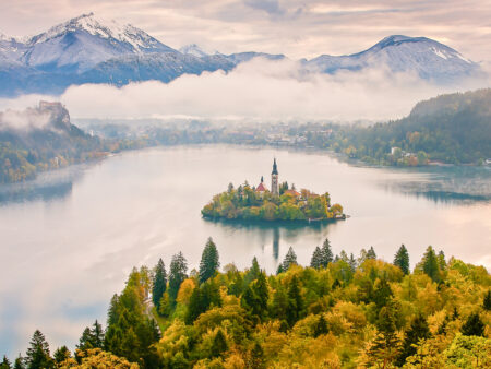 Der Bleder See liegt im Tal vor der Person auf Wanderreise, die das Foto von einer Anhöhe gemacht hat. Im Zentrum des Sees liegt die kleine Insel mit der Kirche darauf. Der Hintergrund ist wolkenverhangen.