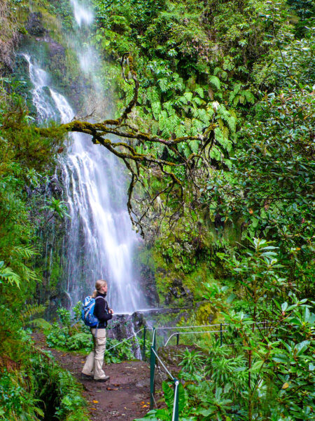Eine Frau mit Wanderrucksack steht vor einem Wasserfall im Erika Lorbeerwald am Beginn des längsten Flusstals Madeiras.