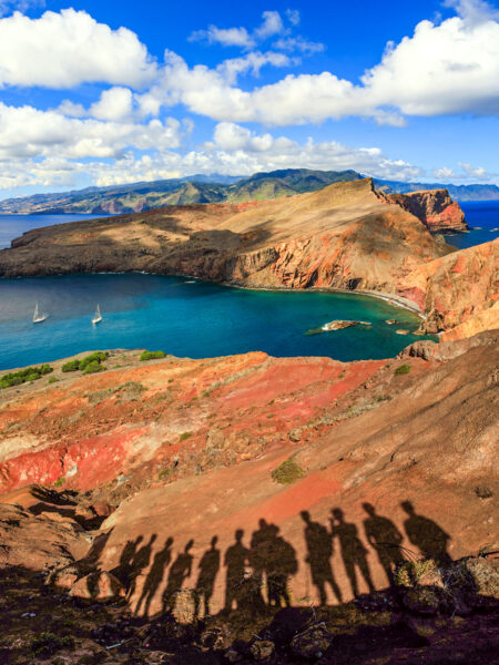 Man sieht die Schatten einer kleinen Gruppe auf Wanderreise auf dem rötlichen Untergrund Madeiras. Im Hintergrund kann man die Küstenlandschaft der Atlantikinsel sehen.