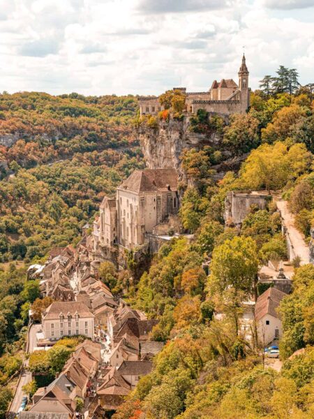 Man sieht die französische Ortschaft Rocamadour in einem herbstlichen Wald. Die alten Gebäude der mittelalterlichen Ortschaft sind an den Felshang gebaut und hängen quasi halb über dem darunter liegenden Tal, das zu Wanderungen durch die herbstliche Landschaft einlädt.