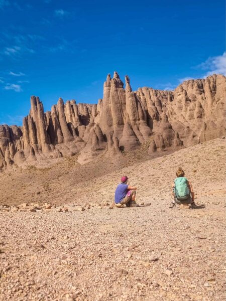 Zwei Personen auf einer Wanderreise durch die Jbel Sarhro sitzen auf dem steinigen Untergrund in Marokko. Im Hintergrund sieht man die spitz zulaufenden Felsformationen des marokkanischen Gebirges.