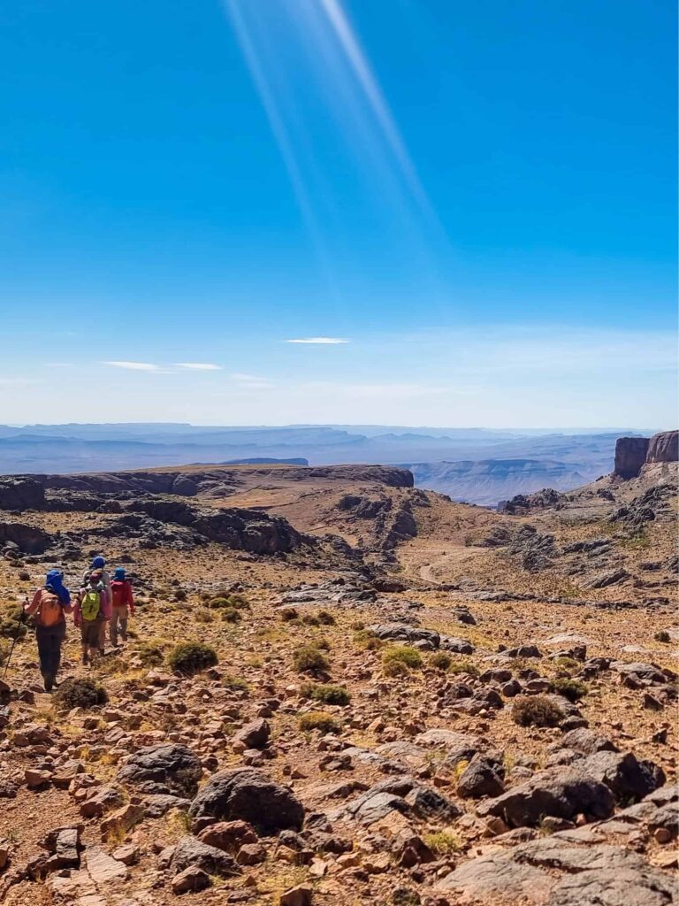 Mehrere Personen wandern durch einen Landstrich in der Jbel Sarhro in Marokko. Sie sind nur klein im Vordrgrund des Bildes zu sehen, während der größte Teil des Fotos von der kargen Gebirgslandschaft eingenommen wird.