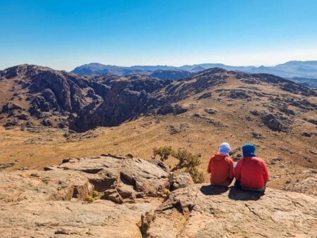 Zwei Wanderreisende sitzen an einer Felskante und schauen hinaus auf die Gebirgslandschaft Marokkos. Sie tragen beide T-Shirts des Reiseanbieters Weltweitwandern.