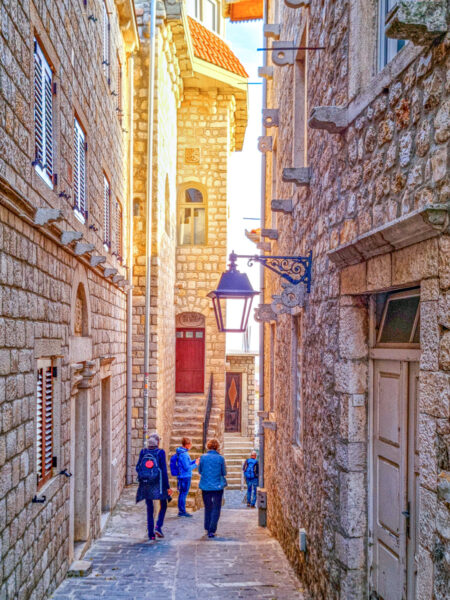Mehrere Personen auf Wanderreise durch Montenegro gehen ein gepflasterte Gasse in Ulcinj hinab. Das Licht der untergehenden Sonne hüllt die Szene in ein warmes Licht.