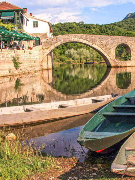 Eine Brück überspannt einen Fluss in der Nähe von Rijeka Crnojevica. Mehrere Ruderboote liegen am Ufer und man sieht eine kleines Restaurant, das Leute auf Wanderreise zu einer erholsamen Pause am Fluss einlädt.