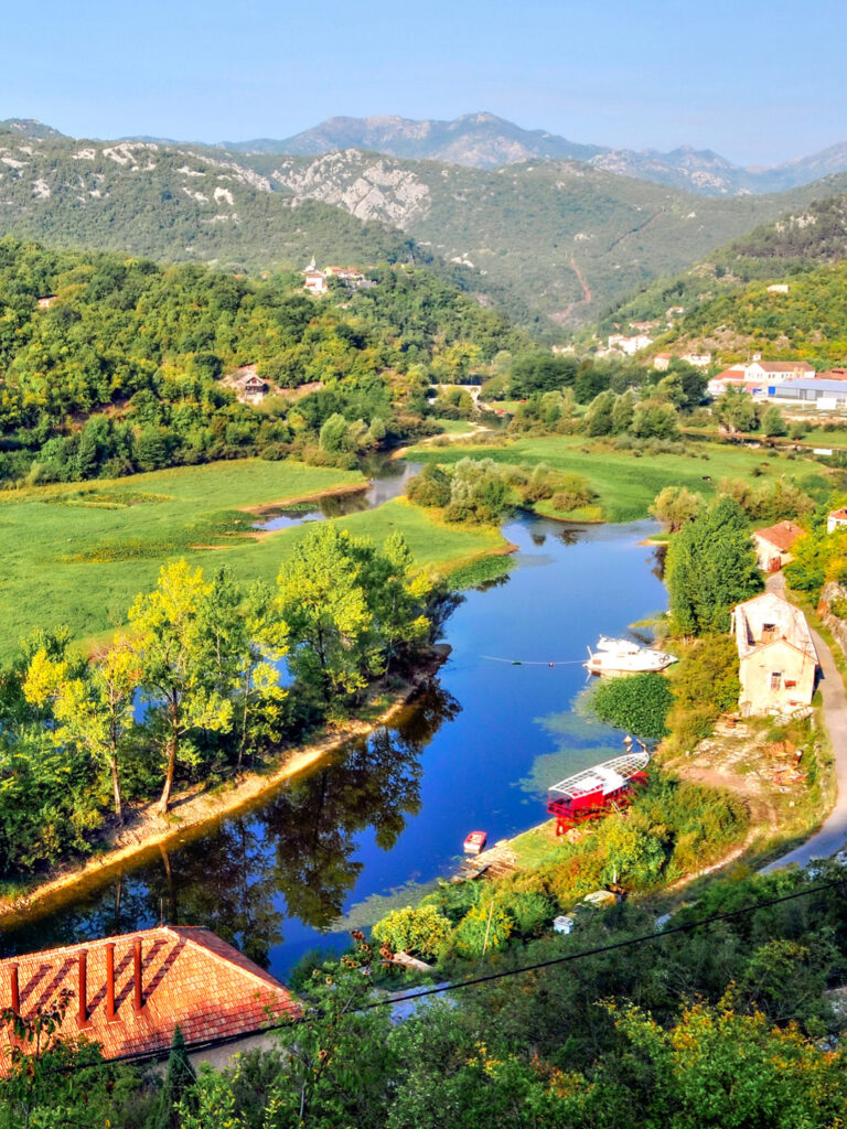 Ein Fluss in der Nähe von Rijeka Crnojevica in Montenegro. Er ist umgeben von grüner Natur und mehrere Gebäude stehen an seinem Flussufer.