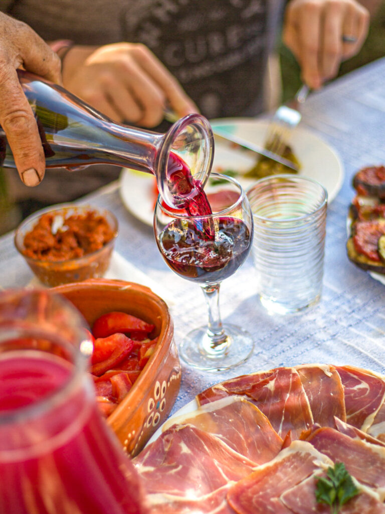 Ein Glas Rotwein wird bei einer kalten Platte in einem Weingut in Montenegro eingeschenkt. Im Hintergrund sieht man eine Person auf Wanderreise, die an der Weinverkostung teilnimmt.