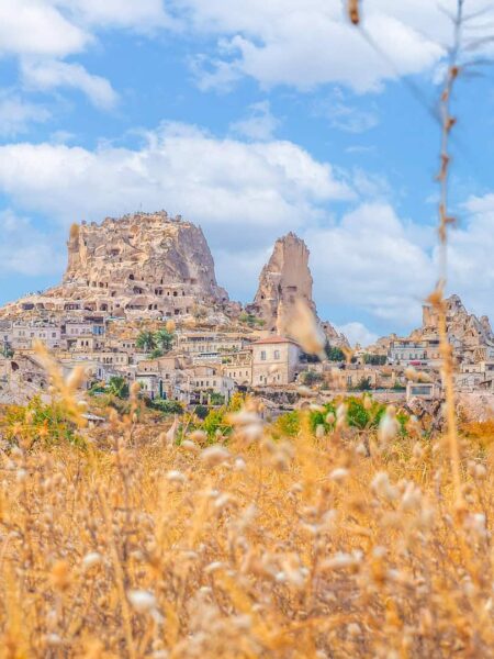 Die Burg Urchisar erhebt sich in der gleichnamigen Stadt üper malerischen Häusern. Im Vordergrund sieht man ein Feld, strahlend blauer Himmel erstreckt sich dahinter.