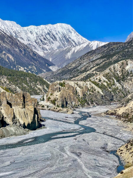 Eine Landschaftsaufnahm im Hochformat entlang des Annapurna-Circuit. Im Zentrum des Bilds fließt ein schmaler Fluss oder Bach durch die Annapurna Conservation Area.