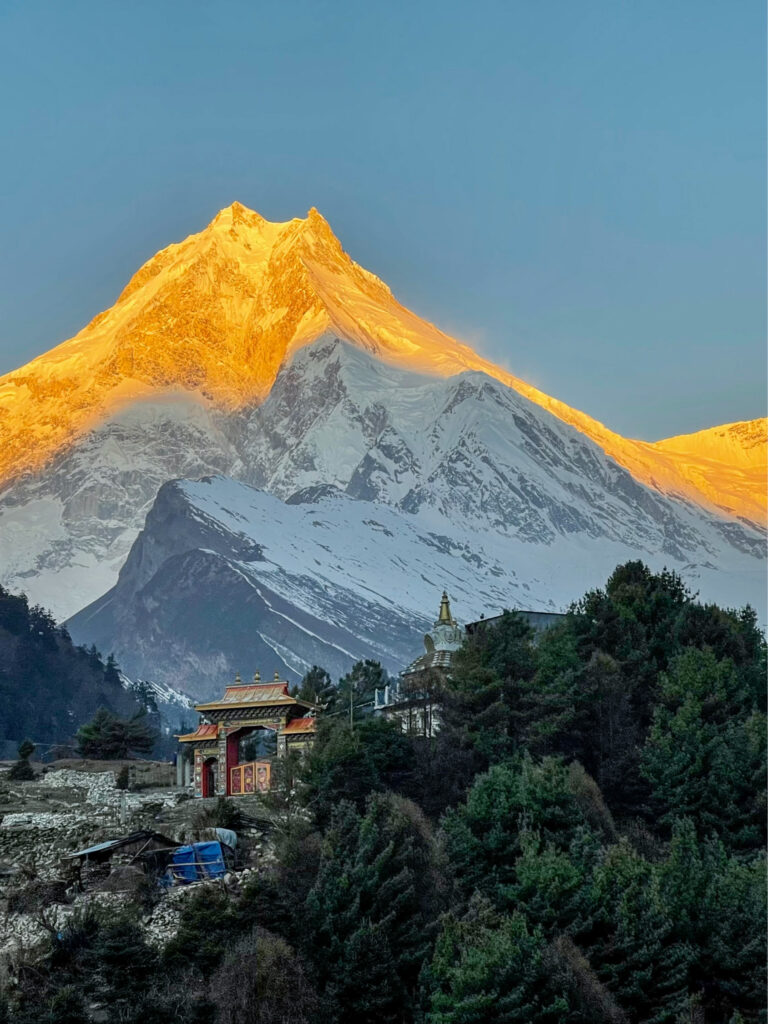 Man sieht den Manaslu bei Sonnenaufgang. Der schneebedeckte Gipfel des Achttausenders ist in warmes Licht gehüllt, während der Rest der Landschaft noch im Schatten liegt.