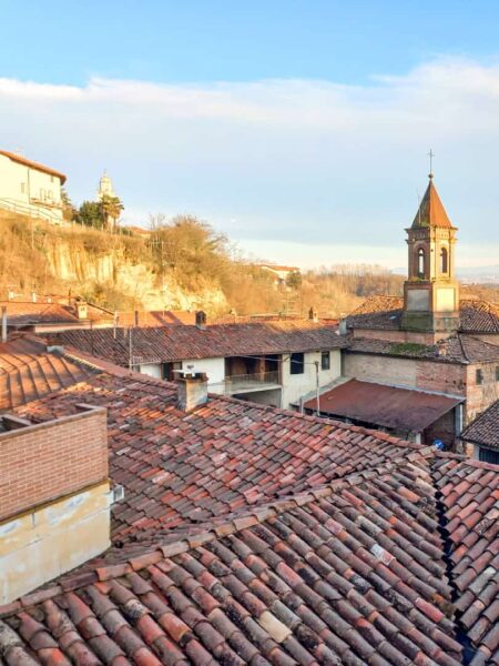 Man kann die roten Dächer einer kleinen italienischen Stadt im Piemont sehen. Das dunkle Rot der Dachziegel hebt sich ab vom Licht der untergehenden Sonne und bietet die perfekte Kulisse für müde Wander-Reisende, um die Beine hochzulegen.