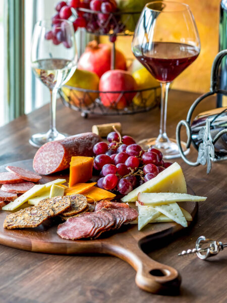 Auf einem Tisch liegt eine kleine Platte mit kalten Köstlichkeiten aus dem Piemont. Man sieht Käse, Wurst, Salami sowie Cracker und Trauben. Zwei Gläser Wein, rot und weiß, steht neben der Jausenplatte.