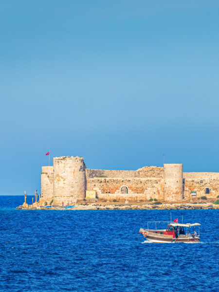Ein Bild der Kızkalesi-Burg an der Südküste der Türkei. Sie liegt direkt am Meer und im Vordergrund fährt ein Boot an der Burg vorbei.