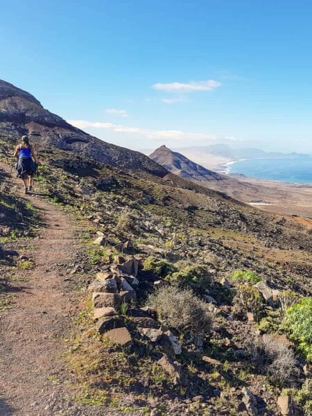 Eine Wanderin folgt einem Pfad auf einer der beiden kanarischen Inseln Lanzarote und Fuerteventura. Im Hintergrund kann man das Meer erkennen.