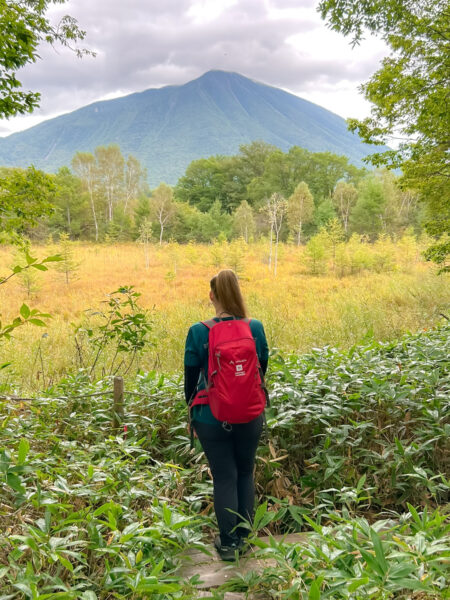 Eine Wanderin mit einem roten Rucksack von Weltweitwandern steht zwischen Büschen neben einem Wanderweg und schaut auf ein Feld hinaus. Im Hintergrund sieht man einen großen Berg in Hokkaido.