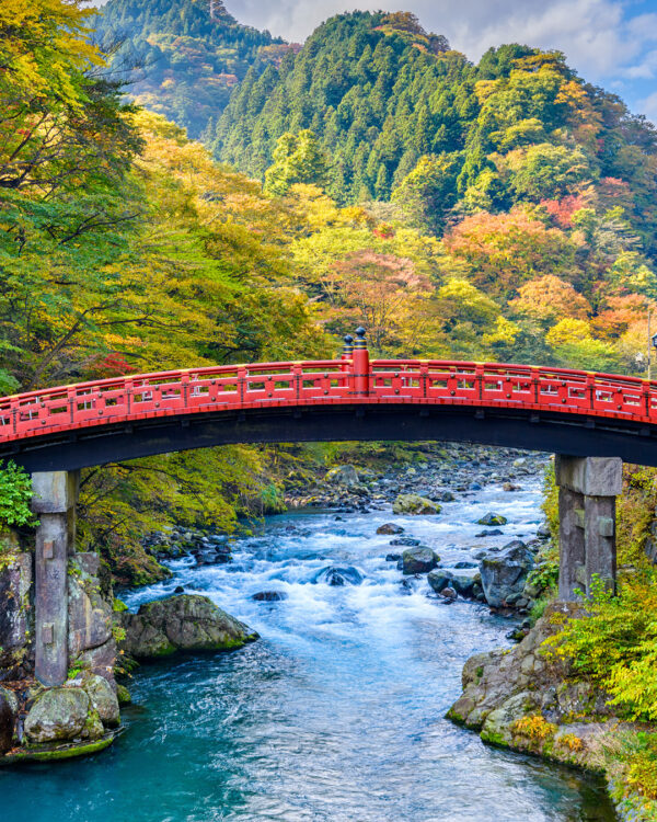 Eine rote Brücke über einen reisenden Fluss im Norden Japans. Im Hintergrund sieht man den herbstlichen Wald von Honshu nahe des Orts Nikko, der nur darauf wartet, während einer Wanderreise erkundet zu werden.