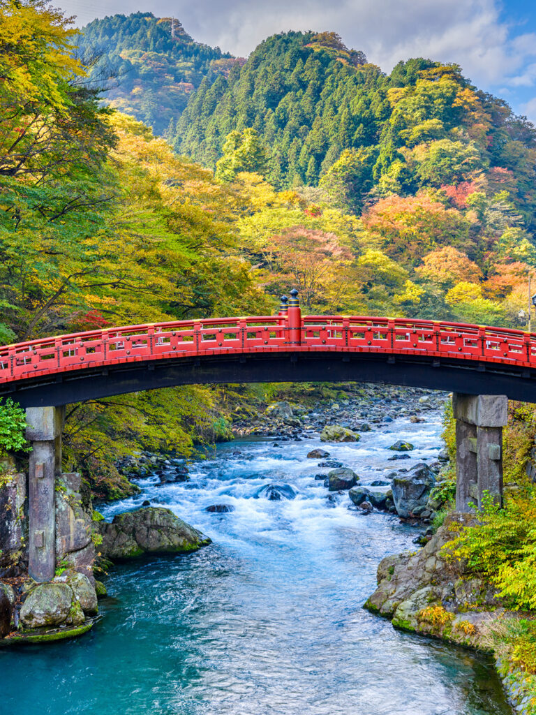 Eine rote Brücke über einen reisenden Fluss im Norden Japans. Im Hintergrund sieht man den herbstlichen Wald von Honshu nahe des Orts Nikko, der nur darauf wartet, während einer Wanderreise erkundet zu werden.