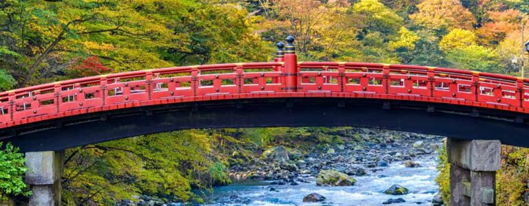 Eine rote Brücke über einen reisenden Fluss im Norden Japans. Im Hintergrund sieht man den herbstlichen Wald von Honshu nahe des Orts Nikko, der nur darauf wartet, während einer Wanderreise erkundet zu werden.