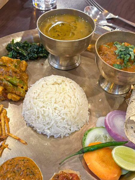Man sieht ein traditionelles Essen aus Nepal auf einem Teller. Gemüse, mehrere Süßen, Süßkartoffeln und ein kleiner Salat sind um Reis herum angerichtet.