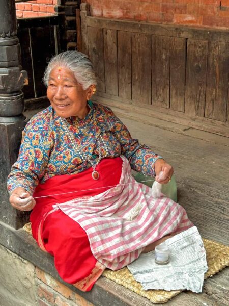 Eine nepalesische Frau aus Bhaktapur sitzt am Boden und lächelt in die Richtung von etwas, das man am Bild nicht erkennen kann. Sie hat graues Haar und trägt bunte Kleidung.