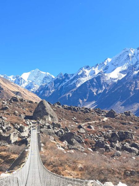 Man sieht eine Hängebrücke aus Metall, die auf ein Bergpanorama in der Region Langtang in Nepal zuführt.