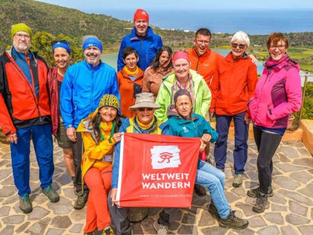 Eine Reisegruppe steht gemeinsam vor einem Ausblick auf Sizilien. Sie halte zwischen sich die Flagge des Reiseveranstalters, mit dem sie hierher auf ihre Wanderreise gekommen sind.
