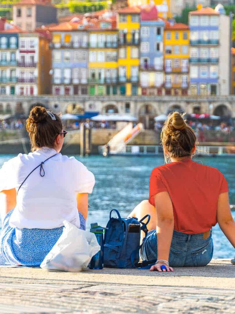 Zwei junge Frauen auf einer Wanderreise mit Weltweitwandern sitzen im Hafen von Porto und schauen hinüber auf die farbenprächtigen Gebäude der Stadt.