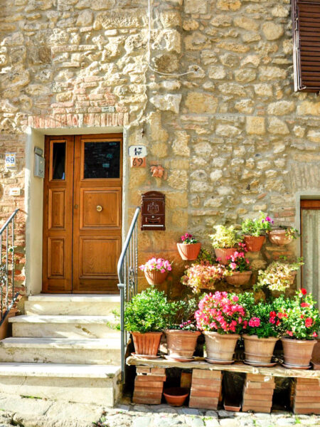 Auf dem Foto kann man Gebäude in einer toskanischen Altstadt sehen. Im Zentrum des Bilds sieht man die Tür des Gebäudes, während davor einige Tröge mit Bluemen und anderen Pflanzen zu sehen sind.