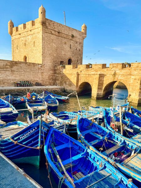 Das Bild zeigt der historischen Hafen von Essouira, einer Stadt im Süden von Marokko. Im Vordergrund liegen blaue Ruderboote im Wasser vertäut, während im Hintergrund die Festung Scala du Port zu sehen ist.