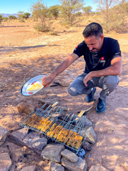 Ein Guide von Weltweitwandern sitzt vor einem kleinen Lagerfeuer mit einem Rost und grillt darauf diverse Spieße.