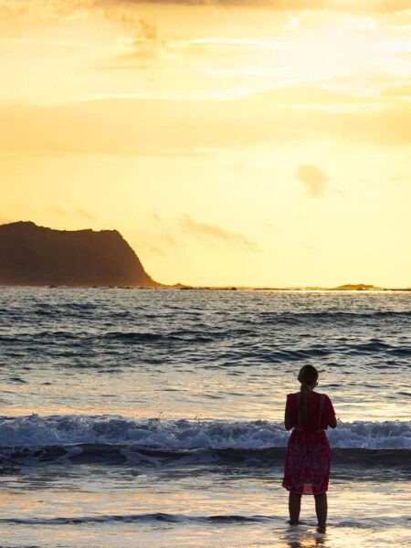 Eine Person auf Wanderreise steht an einem Sandstrand in Costa Rica und schaut hinaus aufs Meer, der untergehenden Sonne entgegen. Die Wellen brechen am weißen Sandstrand.